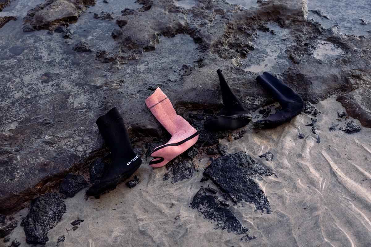 Three pairs of wetsuit booties, two black and one pink, on a rocky surface.