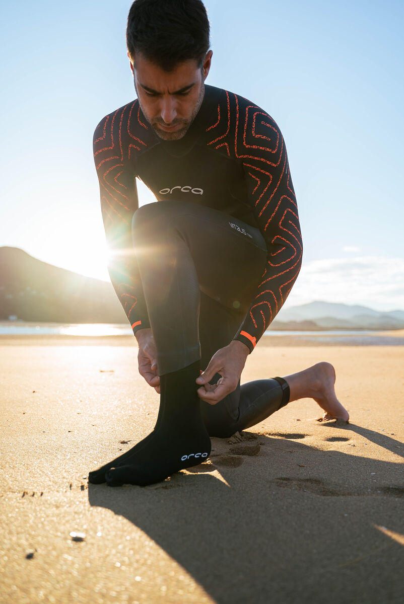 Person wearing an Orca Vitalis wetsuit on a beach with mountains in the background putting on their Hydro swim booties.