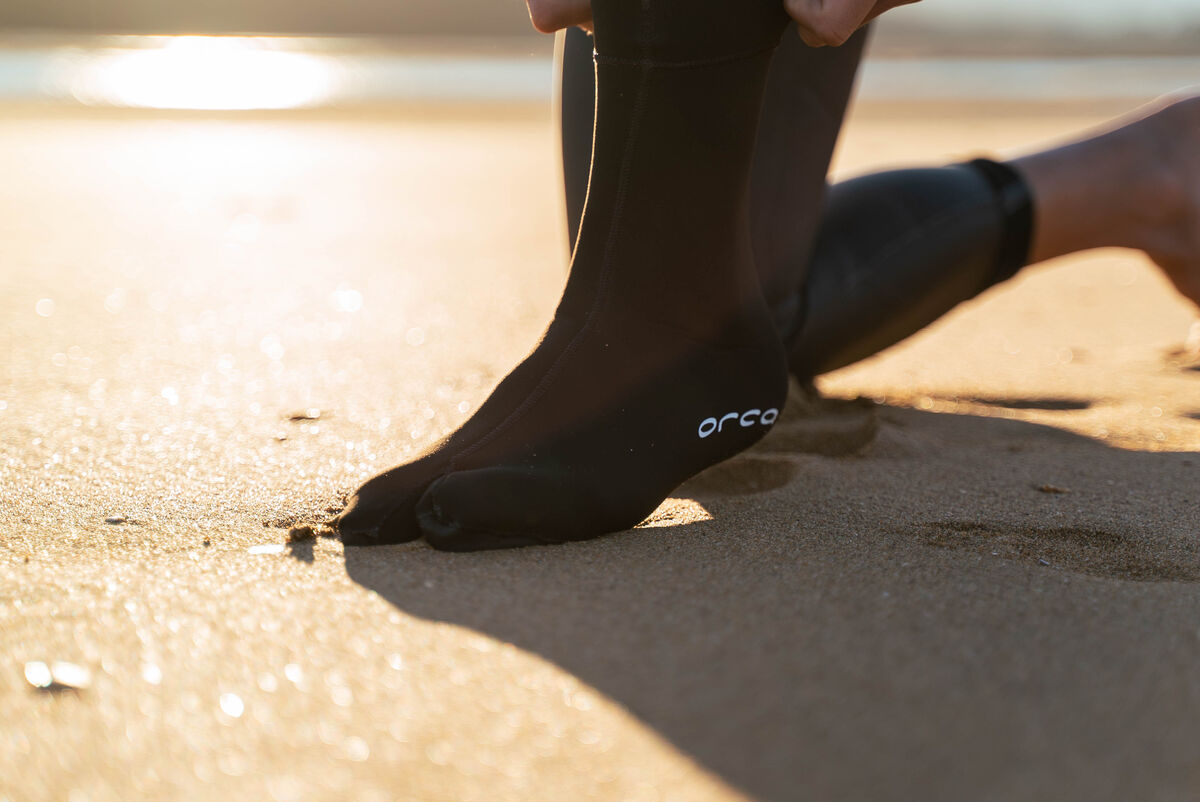 Person wearing black neoprene swim socks with the Orca branding on a sandy surface.