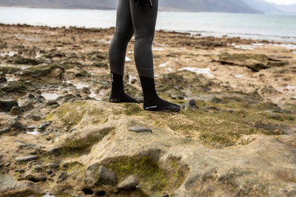 A swimmer wearing their orca hydro booties standing on a rocky shoreline with water and mountains in the background.