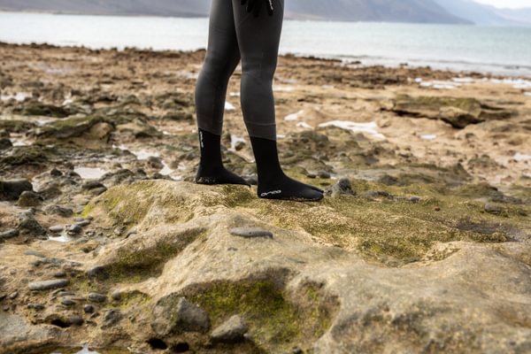 A swimmer wearing their orca hydro booties standing on a rocky shoreline with water and mountains in the background.