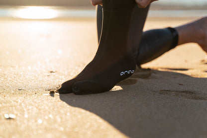 Person wearing black neoprene swim socks with the Orca branding on a sandy surface.